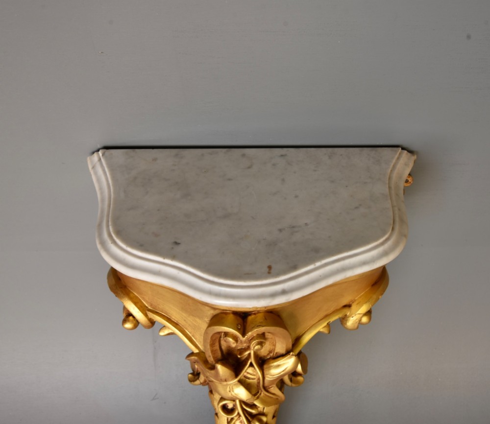 A 19th-century Rococo Revival wall-mounted giltwood console table with a single carved support leg featuring acanthus leaf scrolls and a shaped white marble top. View VI