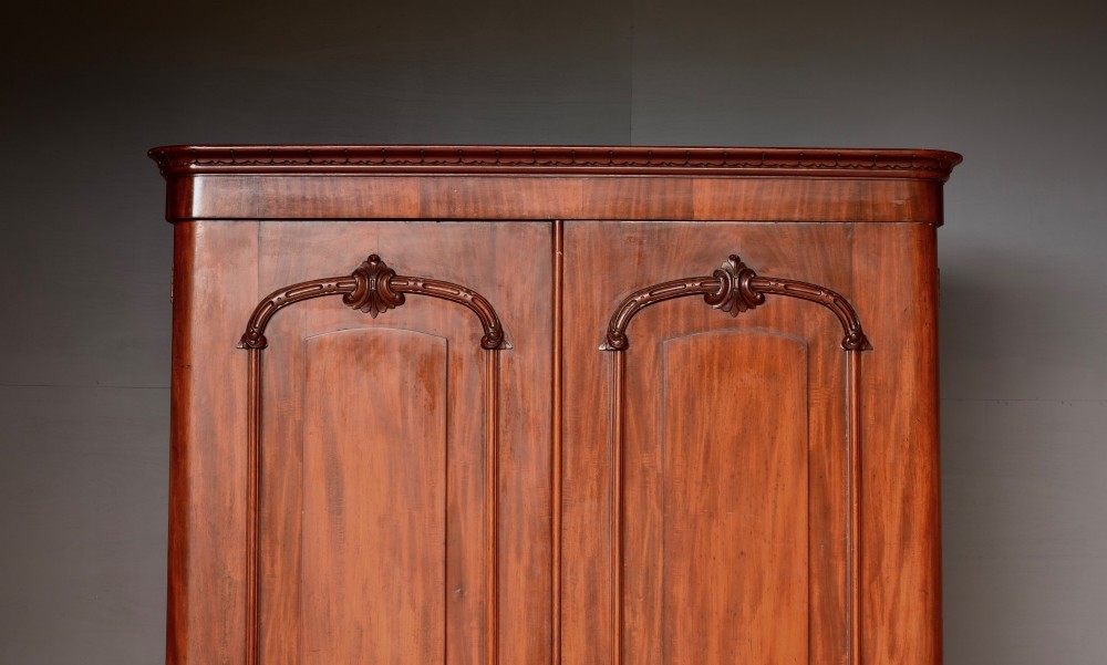 A tall Victorian mahogany double wardrobe with twin panelled doors topped by carved arch mouldings and supported on a plinth base. View II