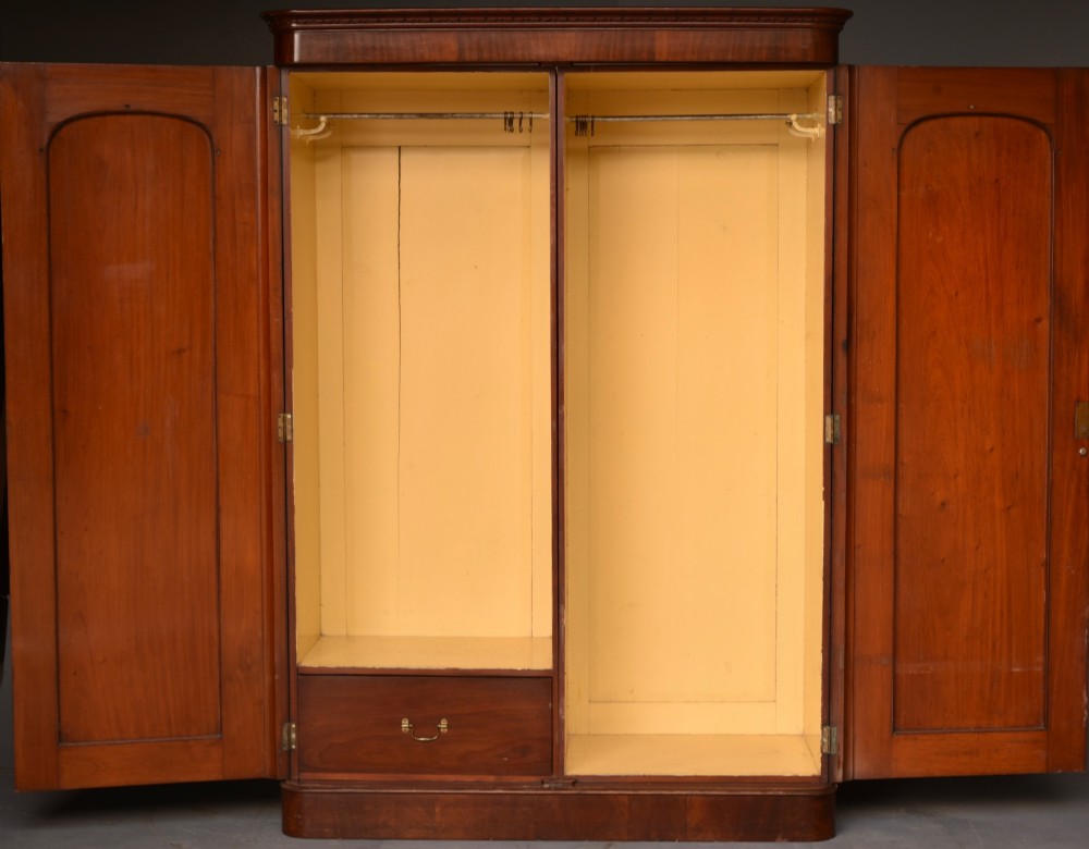 A tall Victorian mahogany double wardrobe with twin panelled doors topped by carved arch mouldings and supported on a plinth base. View V