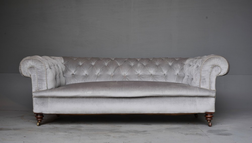A grey velvet Victorian Chesterfield sofa featuring deep-buttoned upholstery, low rolled arms, stud trim, and turned wood legs with brass casters.