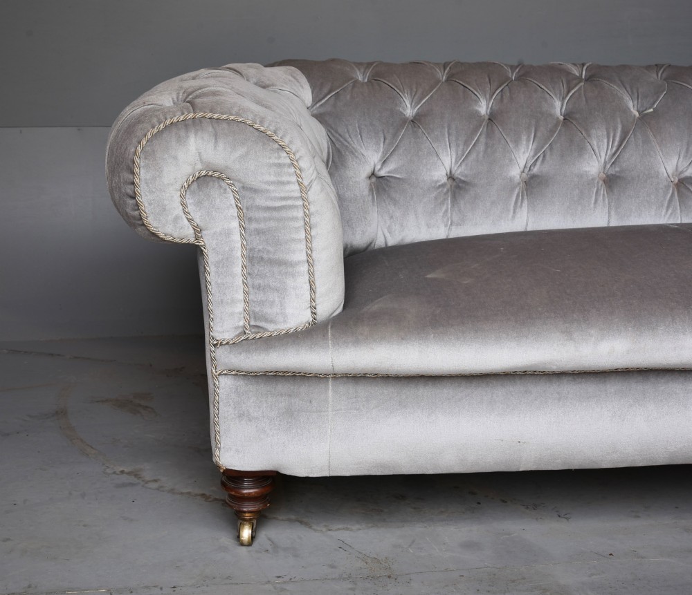 A grey velvet Victorian Chesterfield sofa featuring deep-buttoned upholstery, low rolled arms, stud trim, and turned wood legs with brass casters. View IV