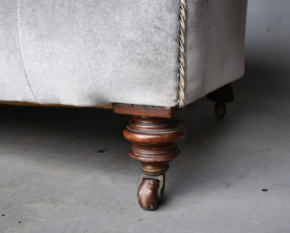 A grey velvet Victorian Chesterfield sofa featuring deep-buttoned upholstery, low rolled arms, stud trim, and turned wood legs with brass casters. View V