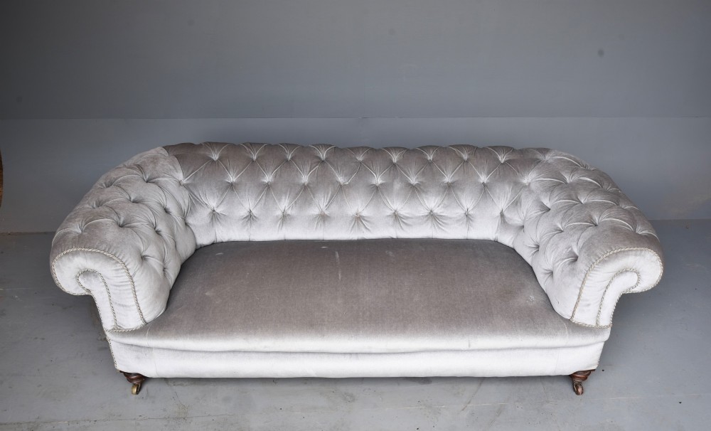A grey velvet Victorian Chesterfield sofa featuring deep-buttoned upholstery, low rolled arms, stud trim, and turned wood legs with brass casters. View VI