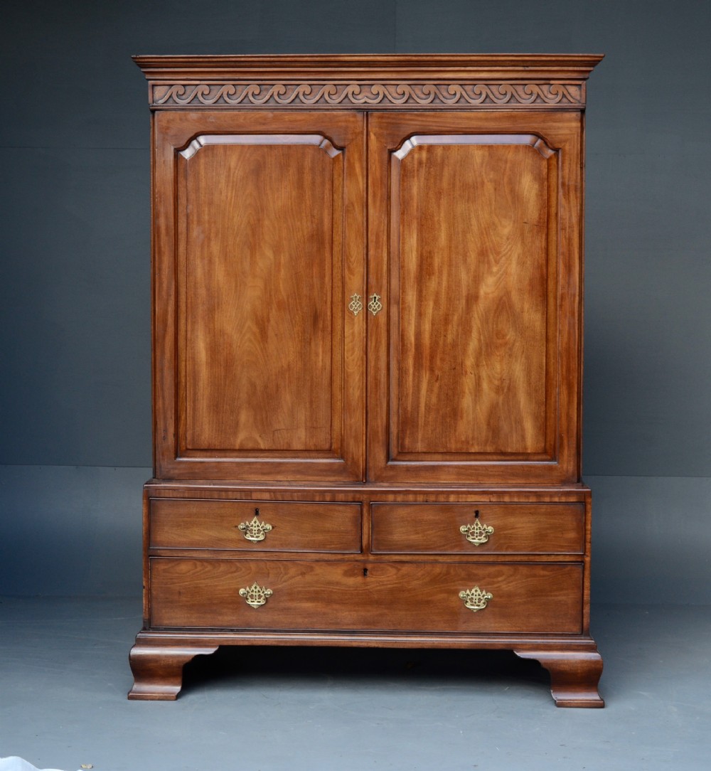 A Georgian mahogany gentlemans wardrobe featuring a carved frieze, double panelled doors, two short over one long drawer, and bracket feet.