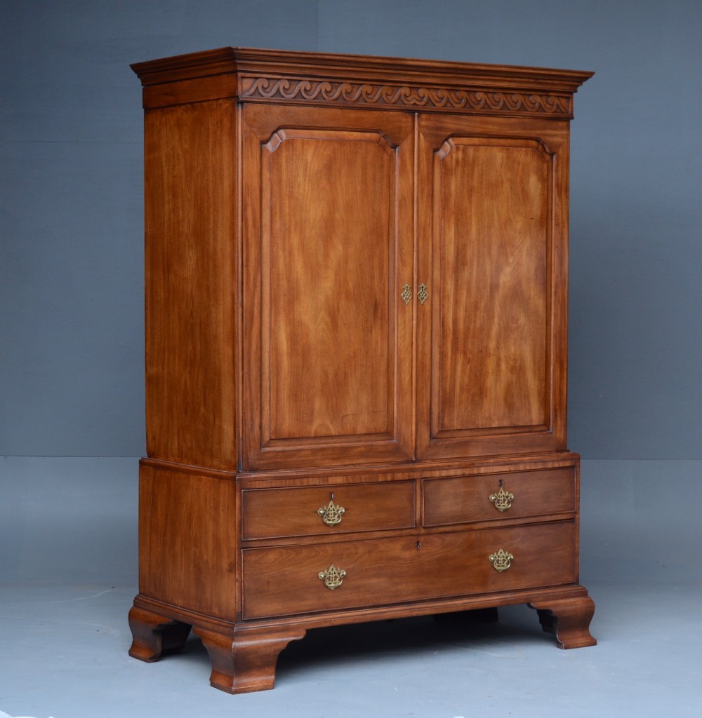 A Georgian mahogany gentlemans wardrobe featuring a carved frieze, double panelled doors, two short over one long drawer, and bracket feet. View II