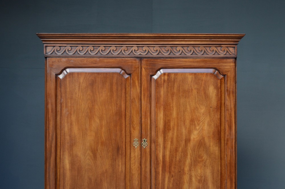 A Georgian mahogany gentlemans wardrobe featuring a carved frieze, double panelled doors, two short over one long drawer, and bracket feet. View IV