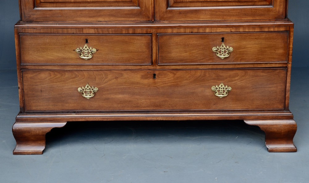 A Georgian mahogany gentlemans wardrobe featuring a carved frieze, double panelled doors, two short over one long drawer, and bracket feet. View V