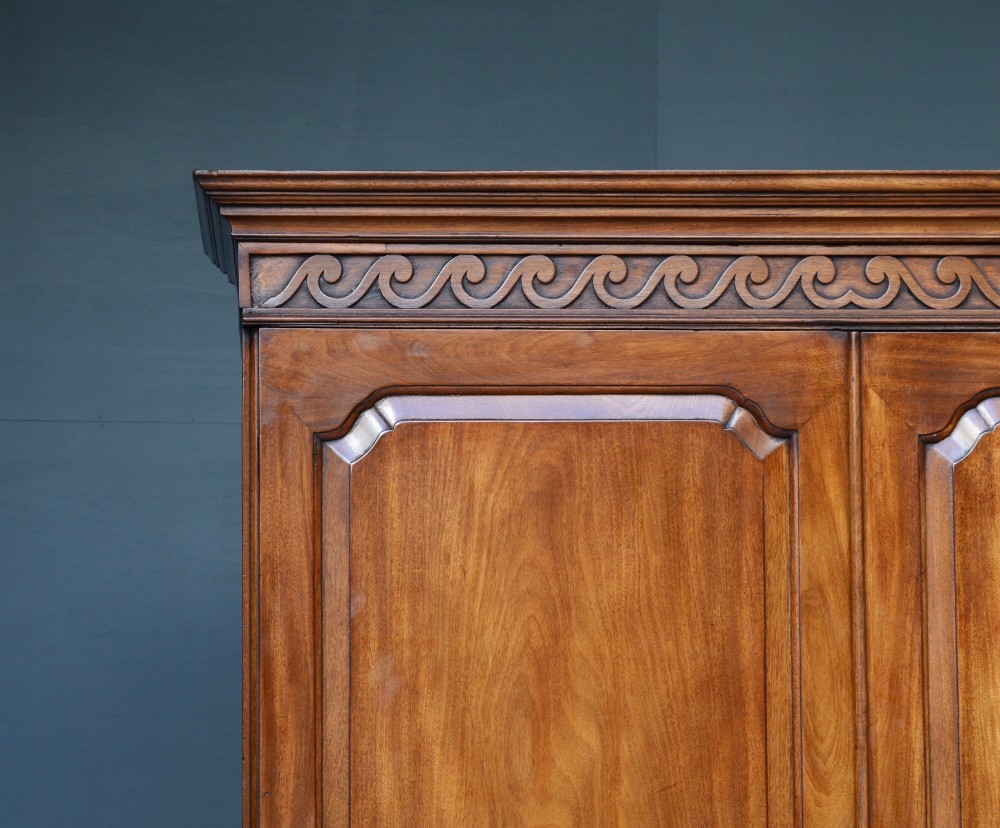 A Georgian mahogany gentlemans wardrobe featuring a carved frieze, double panelled doors, two short over one long drawer, and bracket feet. View VII