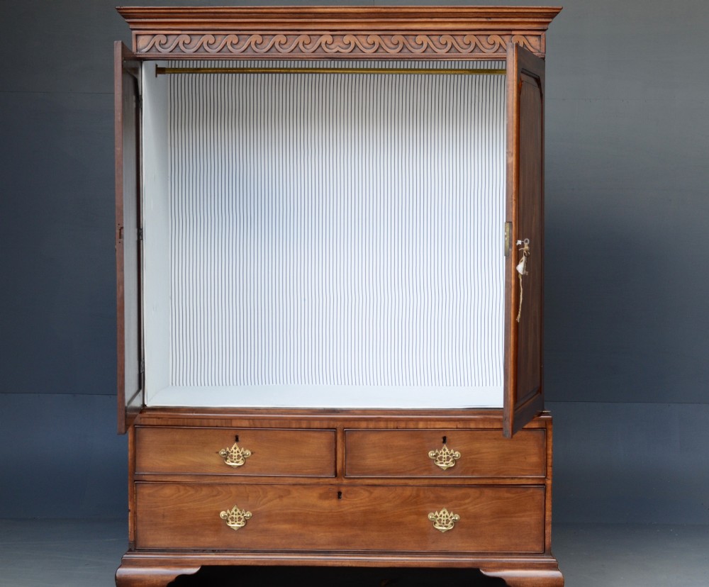 A Georgian mahogany gentlemans wardrobe featuring a carved frieze, double panelled doors, two short over one long drawer, and bracket feet. View VIII