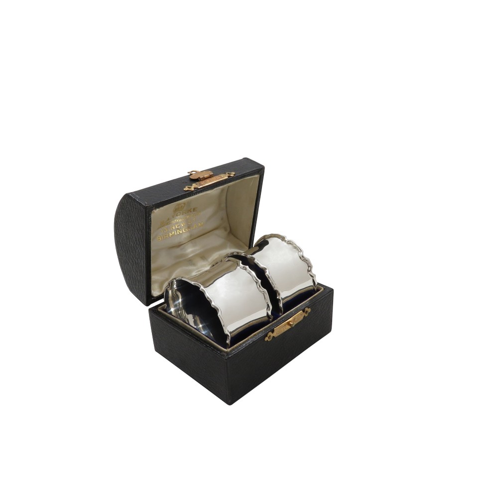 A pair of scalloped sterling silver napkin rings resting inside an open black fitted presentation case lined with cream silk.