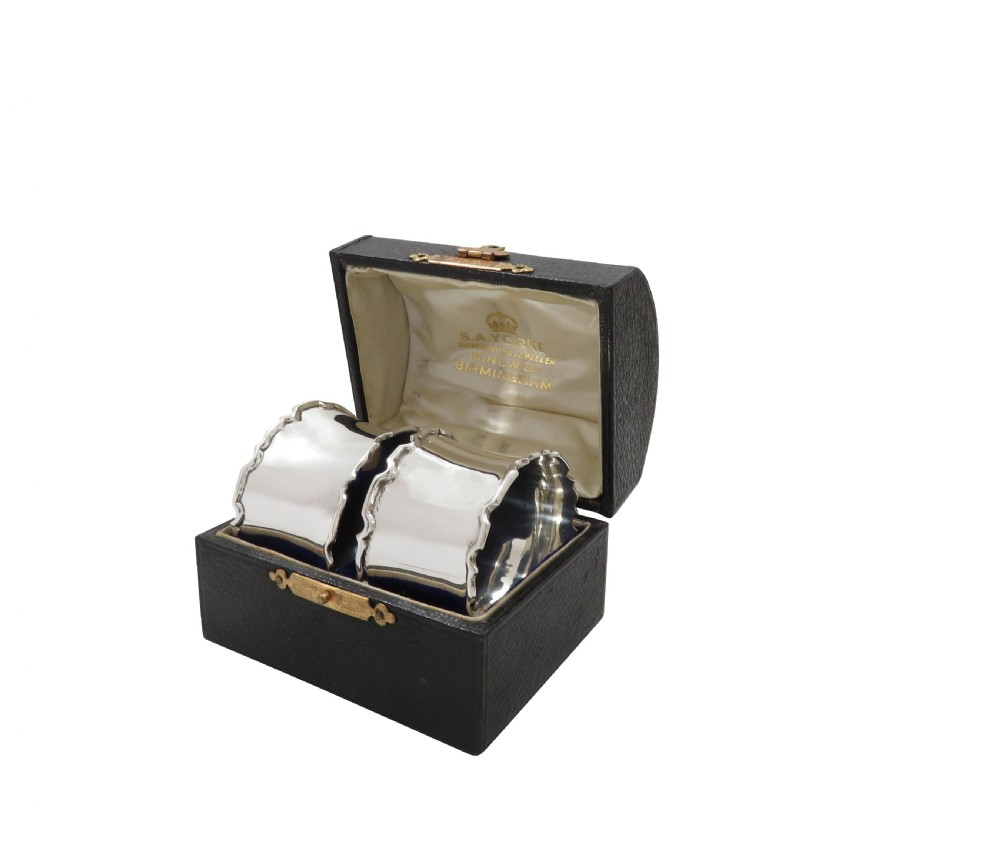 A pair of scalloped sterling silver napkin rings resting inside an open black fitted presentation case lined with cream silk. View IX