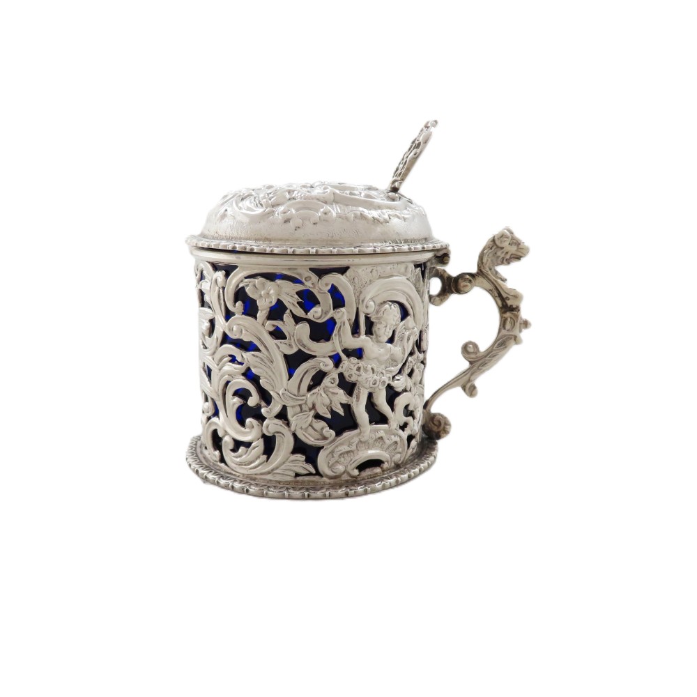 A Victorian sterling silver mustard pot featuring pierced scrollwork and cherub design with a cobalt blue glass liner and matching spoon.