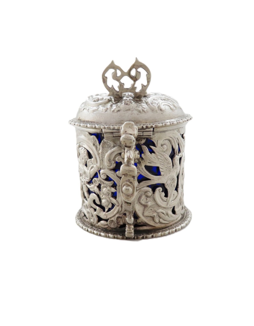 A Victorian sterling silver mustard pot featuring pierced scrollwork and cherub design with a cobalt blue glass liner and matching spoon. View V