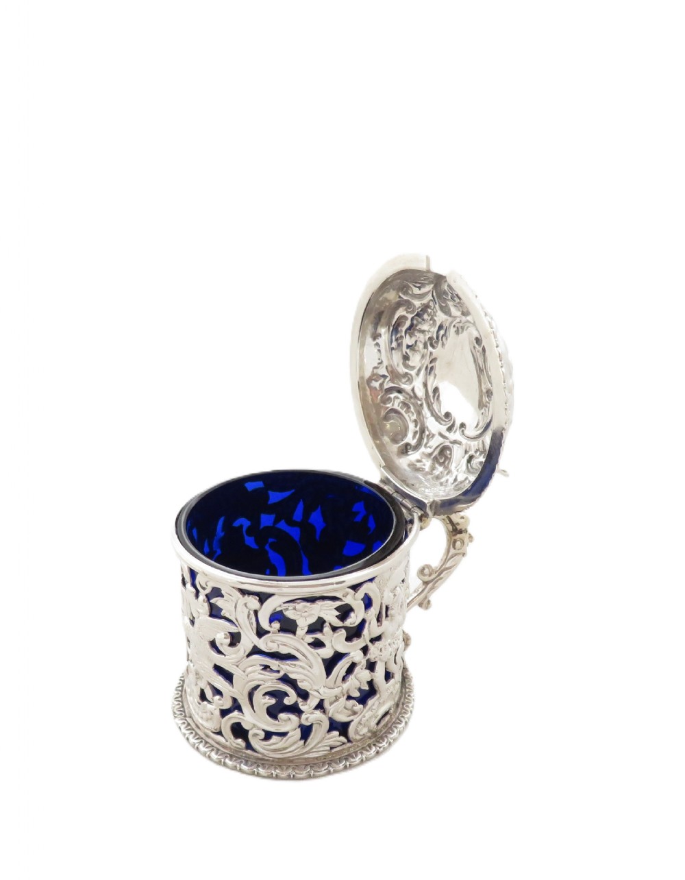 A Victorian sterling silver mustard pot featuring pierced scrollwork and cherub design with a cobalt blue glass liner and matching spoon. View VIII