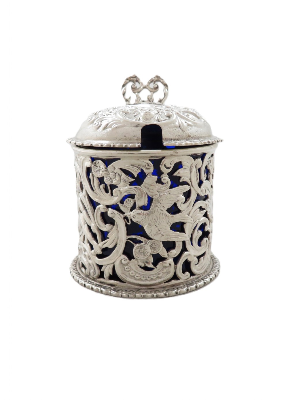 A Victorian sterling silver mustard pot featuring pierced scrollwork and cherub design with a cobalt blue glass liner and matching spoon. View IX