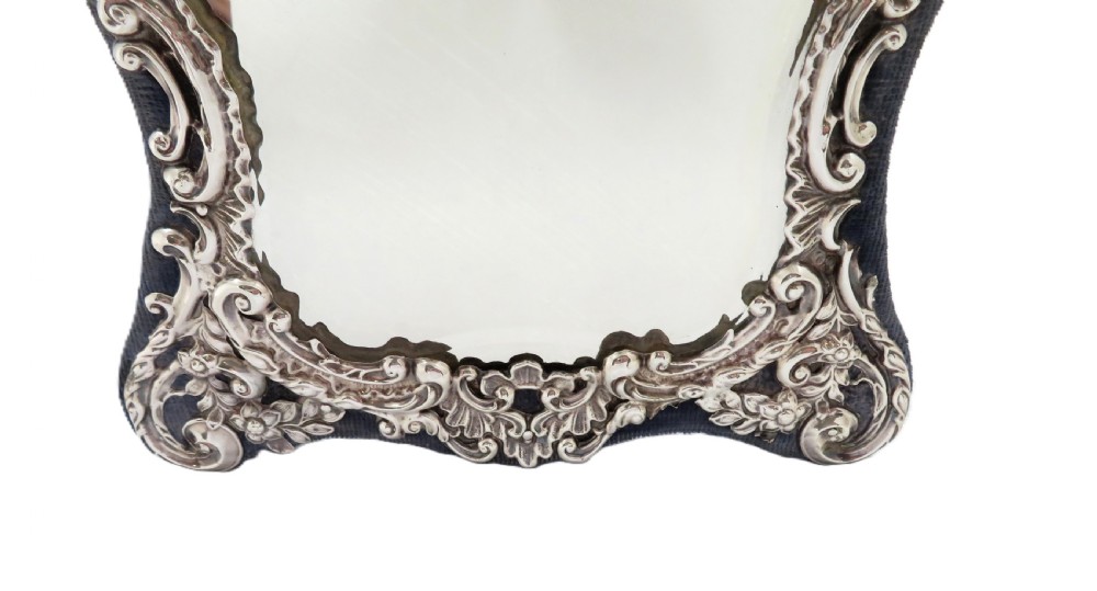 A Victorian sterling silver easel mirror featuring intricate floral repousse decoration and a dark blue velvet backing. View VI