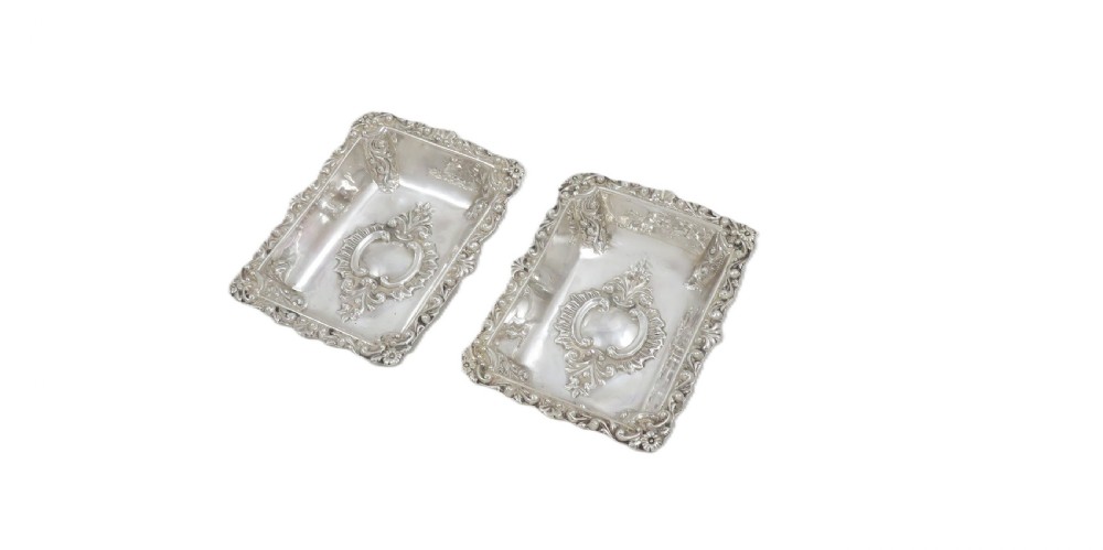A pair of rectangular embossed sterling silver dishes with ornate scrolled borders, displayed inside a silk and velvet-lined dark presentation case. View VI