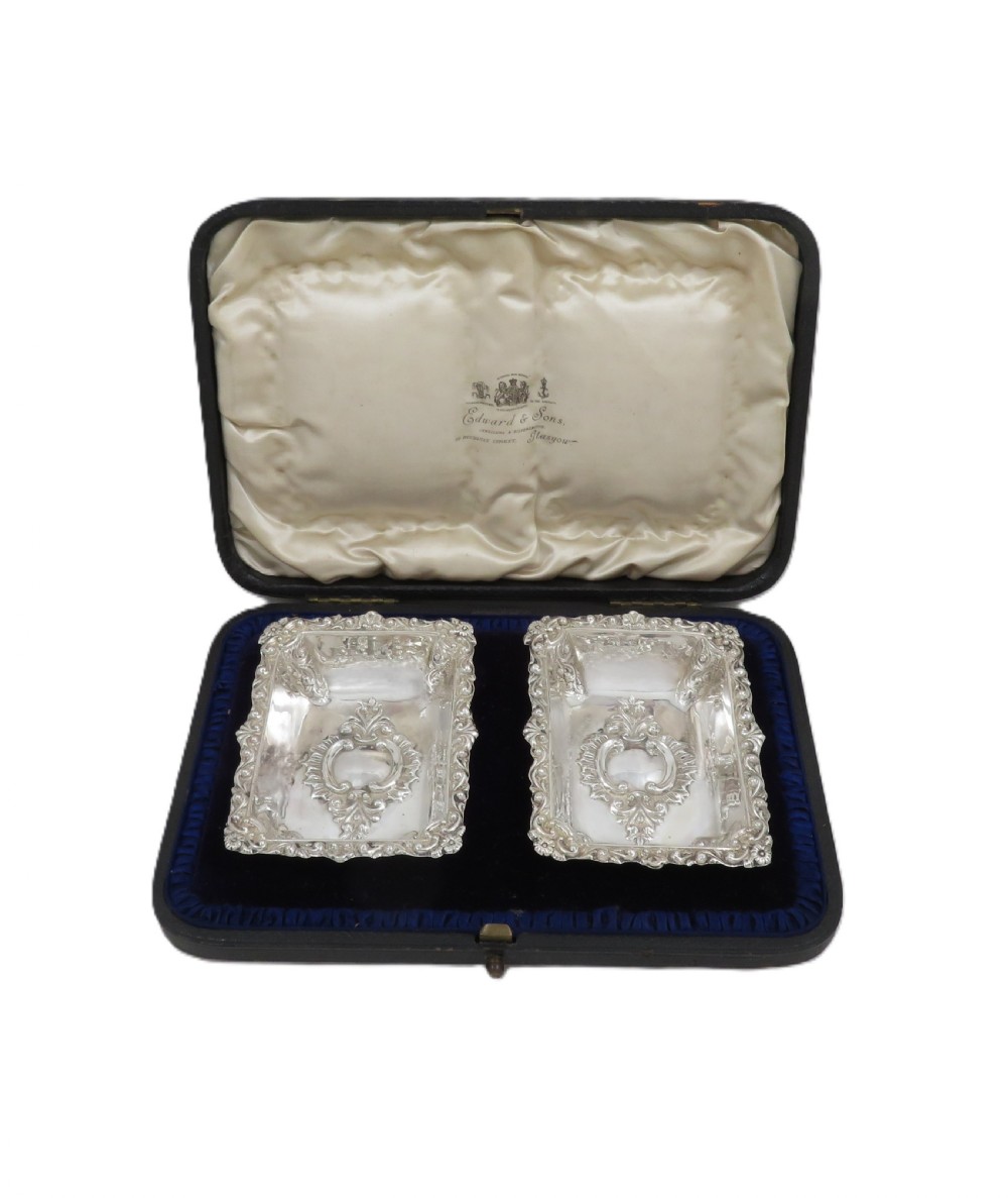A pair of rectangular embossed sterling silver dishes with ornate scrolled borders, displayed inside a silk and velvet-lined dark presentation case. View VII