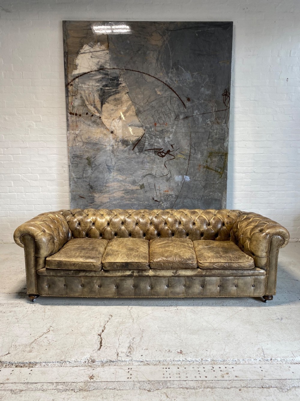 A large mid-century four-cushion Chesterfield sofa upholstered in distressed tan leather with deep button tufting, rolled arms, and brass stud trim.
