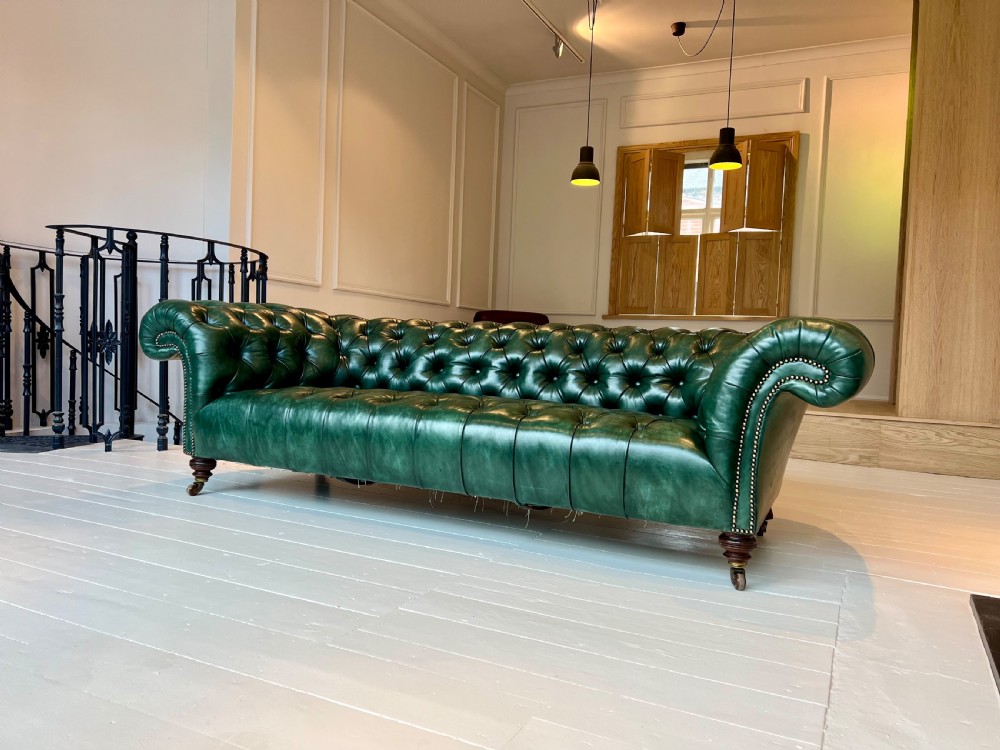 A nineteenth-century green leather Chesterfield sofa featuring deep button tufting, rolled arms with brass nailhead trim, and turned mahogany legs on brass castors. View II
