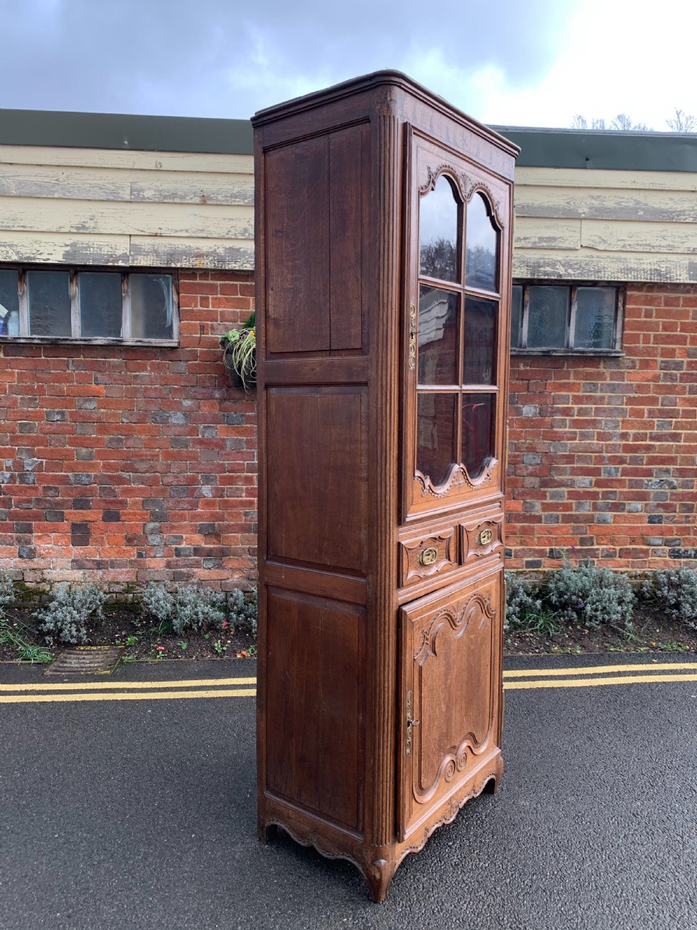 A tall, narrow antique oak cabinet with a glazed upper door, central carved drawers, and a lower panelled cupboard door. View VI