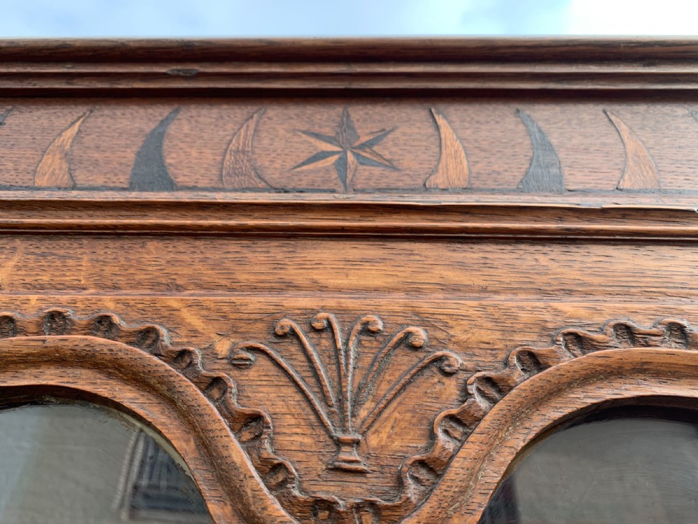 A tall, narrow antique oak cabinet with a glazed upper door, central carved drawers, and a lower panelled cupboard door. View XI