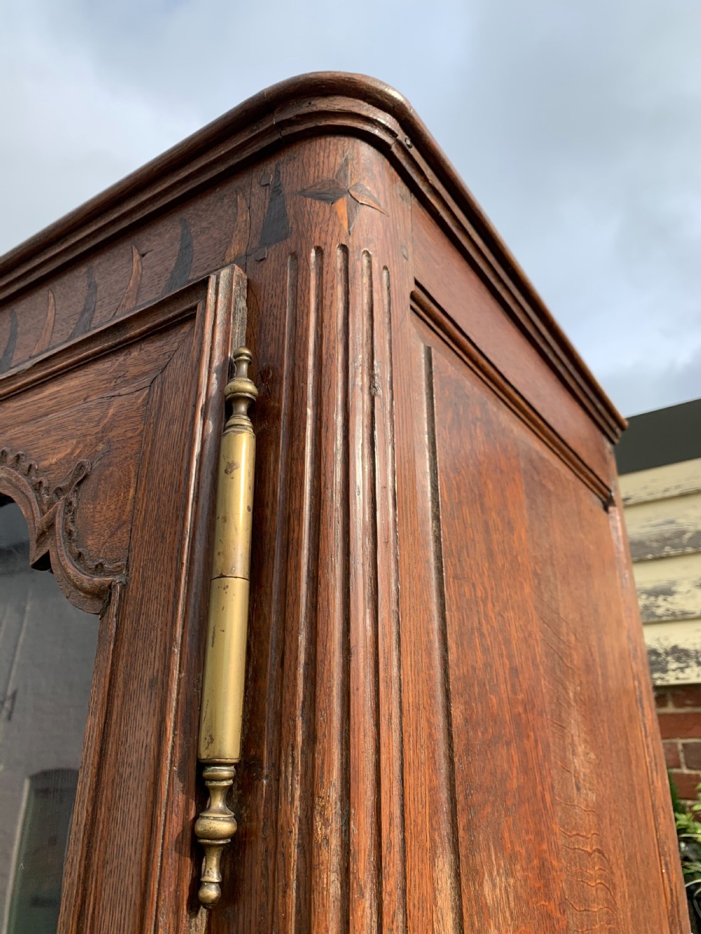 A tall, narrow antique oak cabinet with a glazed upper door, central carved drawers, and a lower panelled cupboard door. View XII
