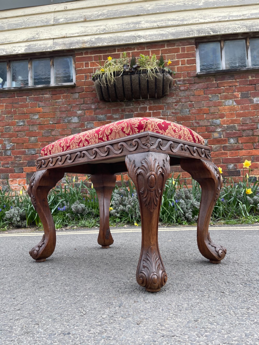 A carved teak Anglo-Indian stool featuring ornate cabriole legs, a decorative carved apron, and a red damask upholstered top.