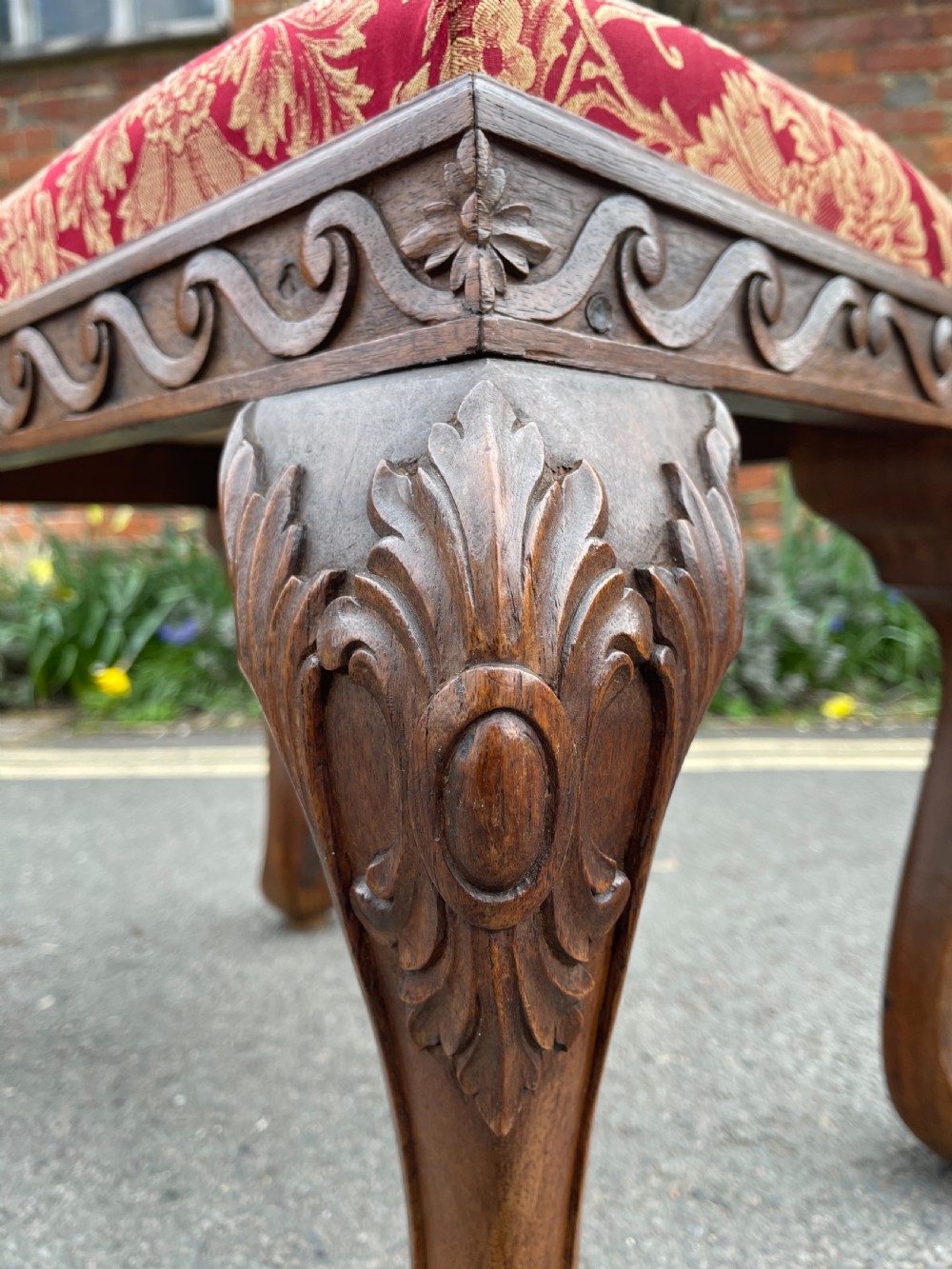 A carved teak Anglo-Indian stool featuring ornate cabriole legs, a decorative carved apron, and a red damask upholstered top. View II
