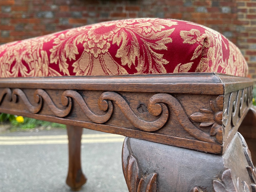 A carved teak Anglo-Indian stool featuring ornate cabriole legs, a decorative carved apron, and a red damask upholstered top. View VII