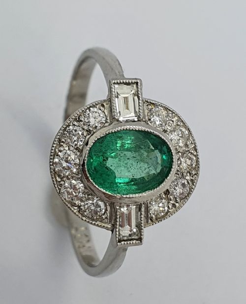 fig 1. An Art Deco style white metal ring featuring a central oval green emerald within a circular diamond halo accented by baguette-cut side diamonds.