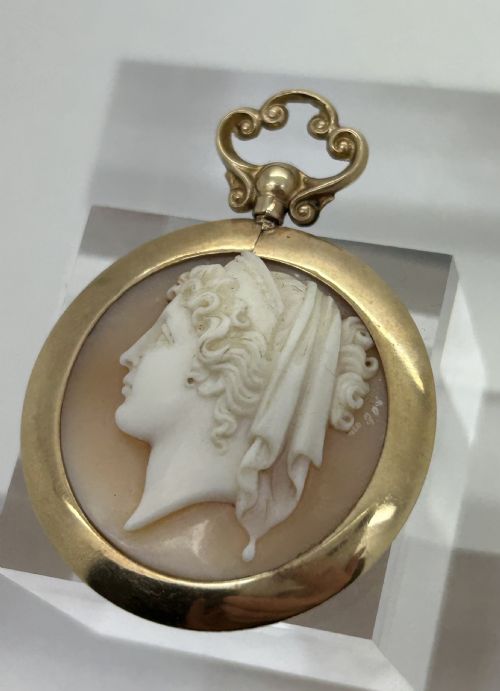 fig 1. An antique round shell cameo depicting a carved female classical profile in cream set against a tan background, encased in a smooth gold frame with a top scroll bail.