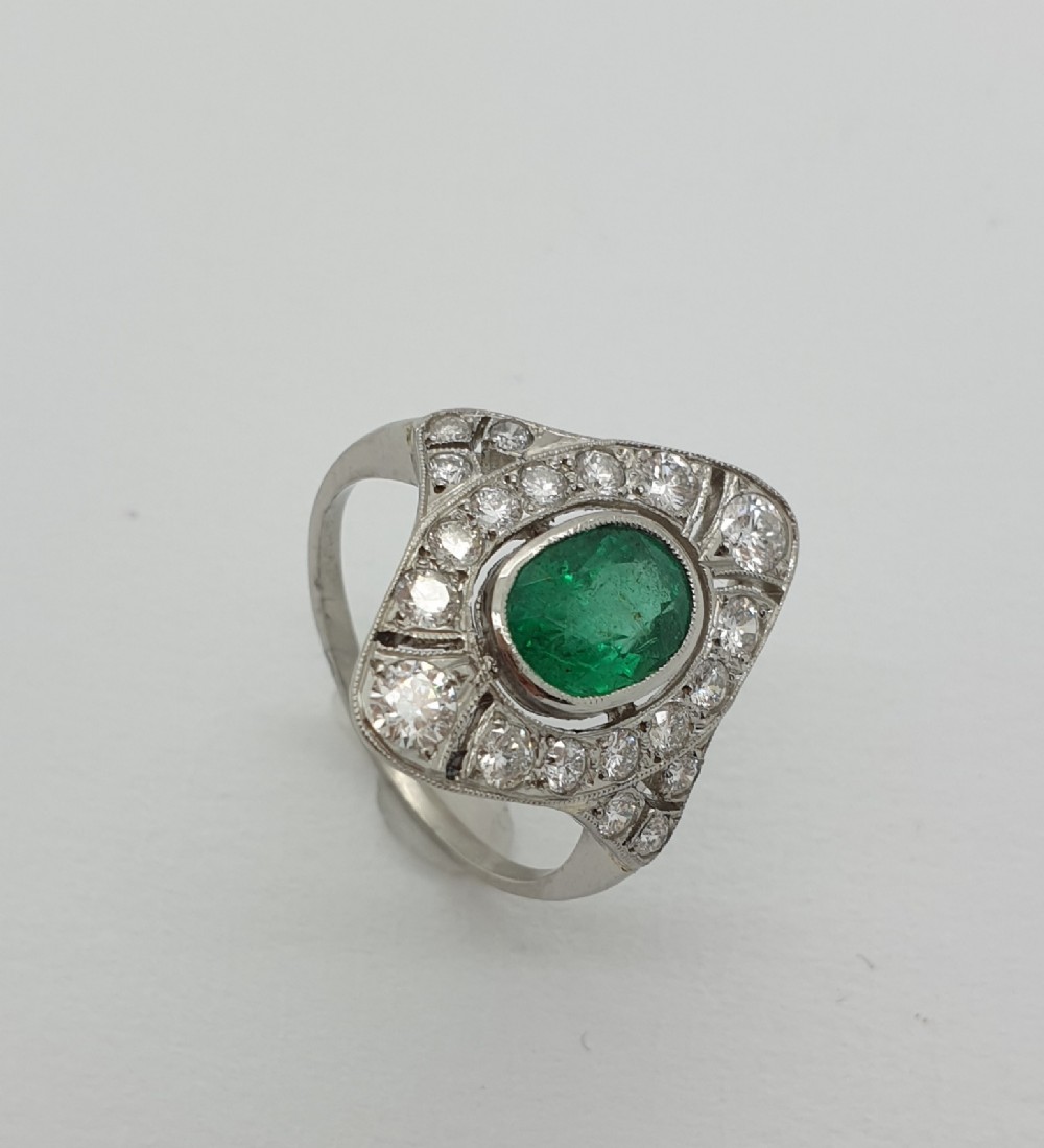 An Art Deco style 18ct white gold cluster ring featuring a central oval emerald framed by a geometric panel of brilliant diamonds with milgrain detailing.