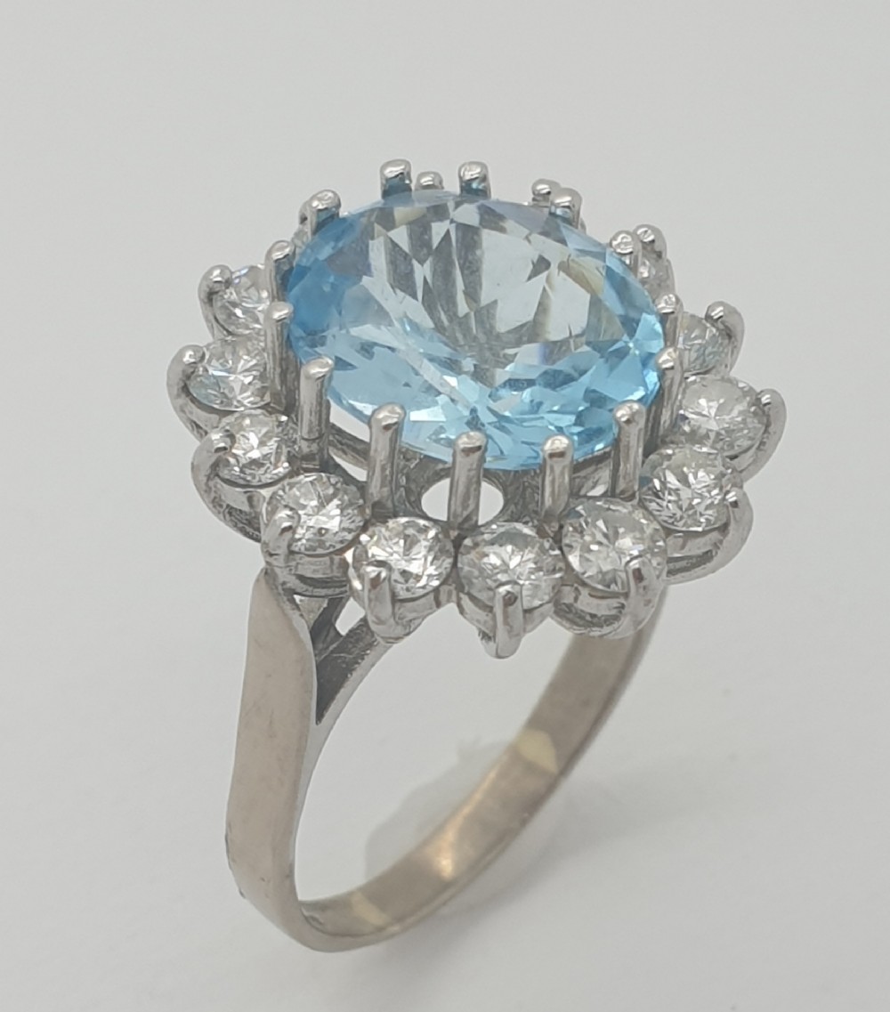 An oval light blue aquamarine central gemstone surrounded by a halo of round brilliant diamonds set in a clawed white metal band.