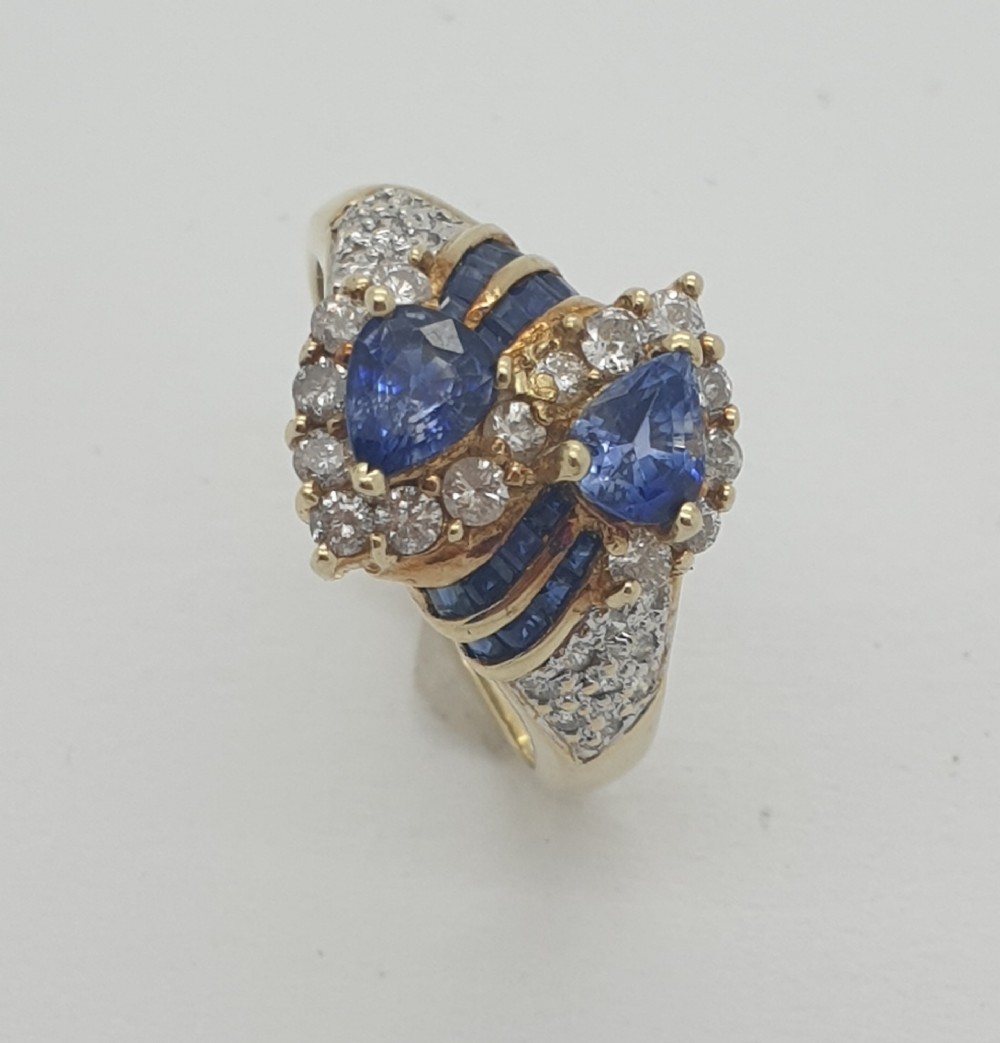 A 14ct yellow gold dress ring featuring two pear-shaped blue tanzanites surrounded by halo diamonds and channel-set accent stones.