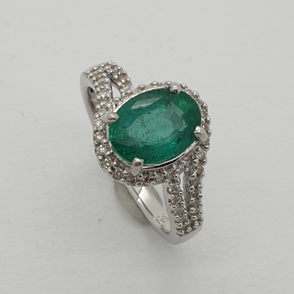 An 18ct white gold ring featuring an oval-cut central green emerald framed by small diamonds extending down the split band.