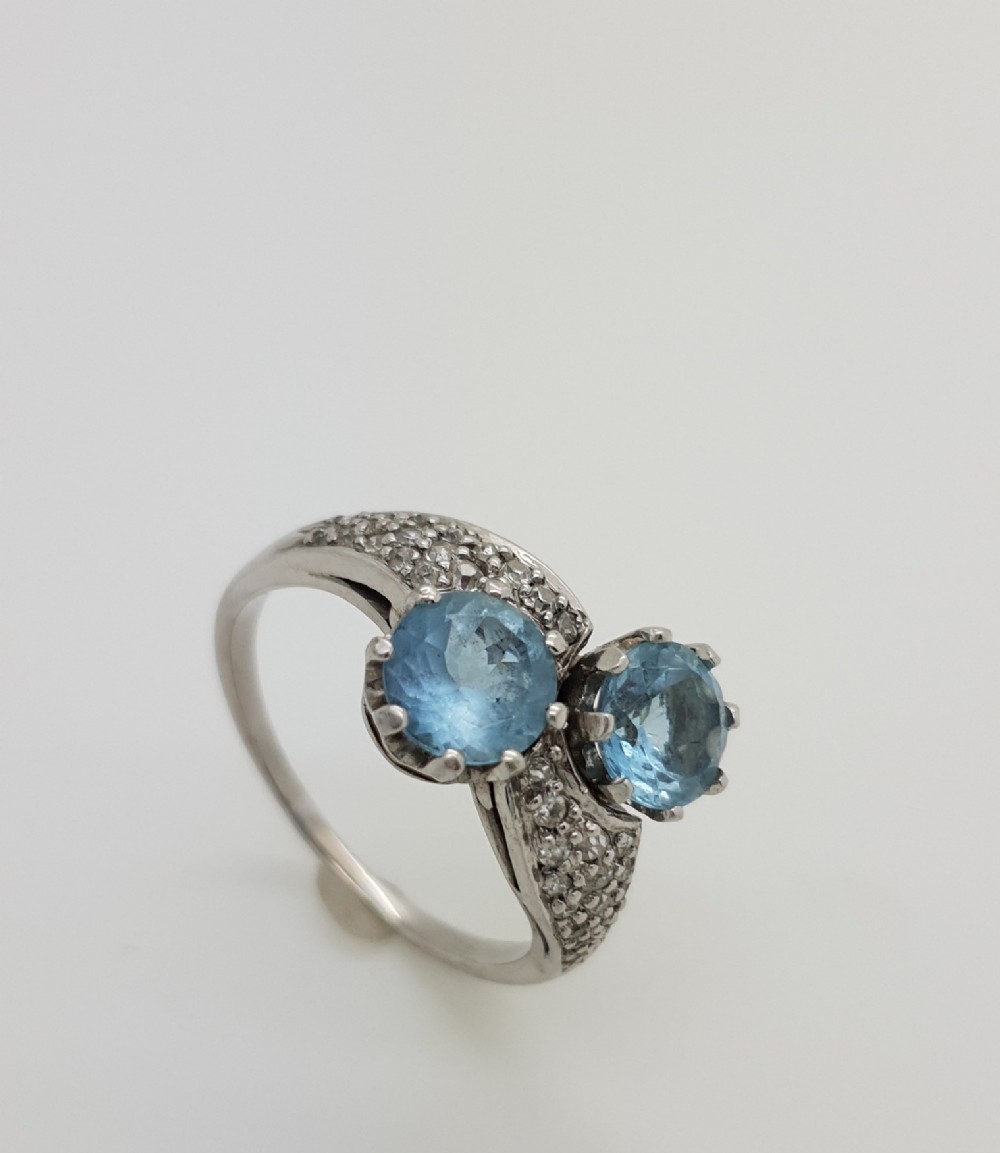 A white metal twist ring set with two round light blue aquamarines and small accent diamonds along the shoulders.