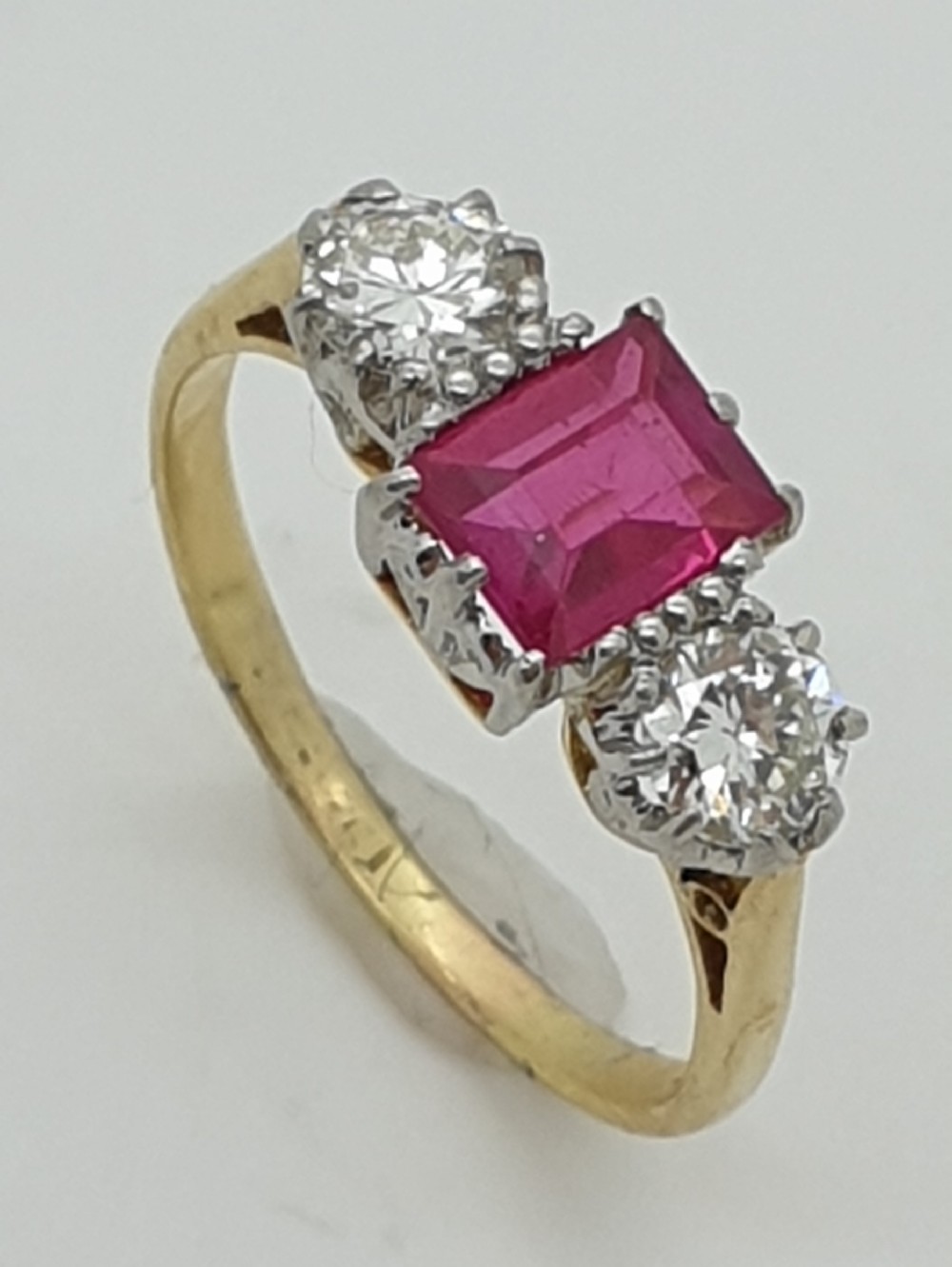 A yellow gold trilogy ring featuring a central rectangular cut red ruby flanked by two round diamonds set in white metal claws.
