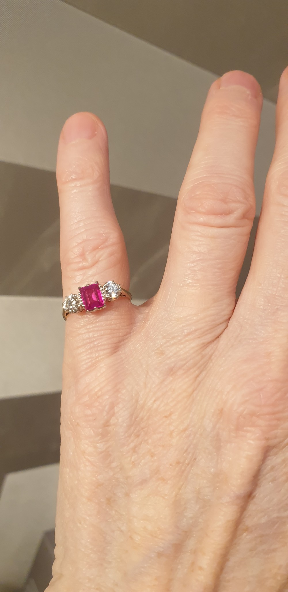 A yellow gold trilogy ring featuring a central rectangular cut red ruby flanked by two round diamonds set in white metal claws. View V