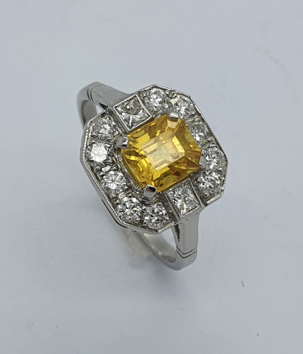 An Art Deco style platinum ring featuring a central emerald-cut yellow sapphire enclosed within an octagonal halo of brilliant and square cut diamonds.
