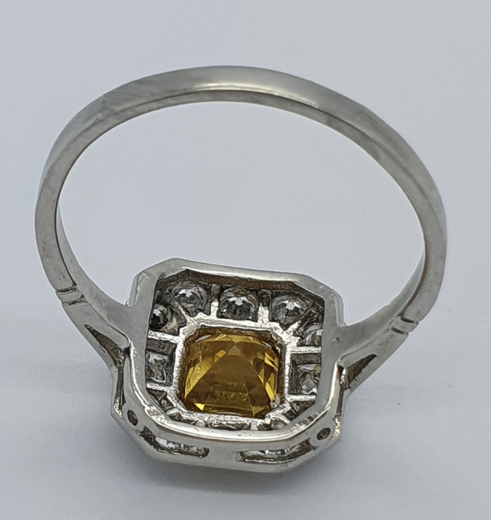 An Art Deco style platinum ring featuring a central emerald-cut yellow sapphire enclosed within an octagonal halo of brilliant and square cut diamonds. View IV