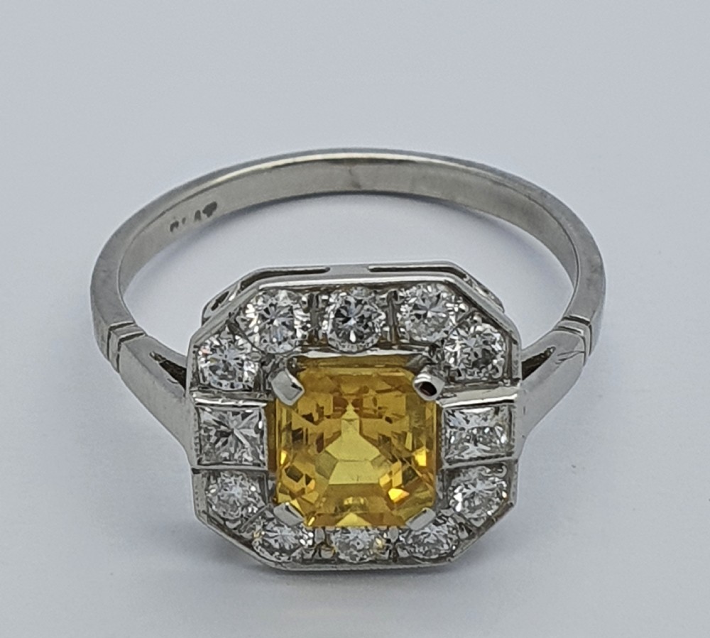 An Art Deco style platinum ring featuring a central emerald-cut yellow sapphire enclosed within an octagonal halo of brilliant and square cut diamonds. View VI