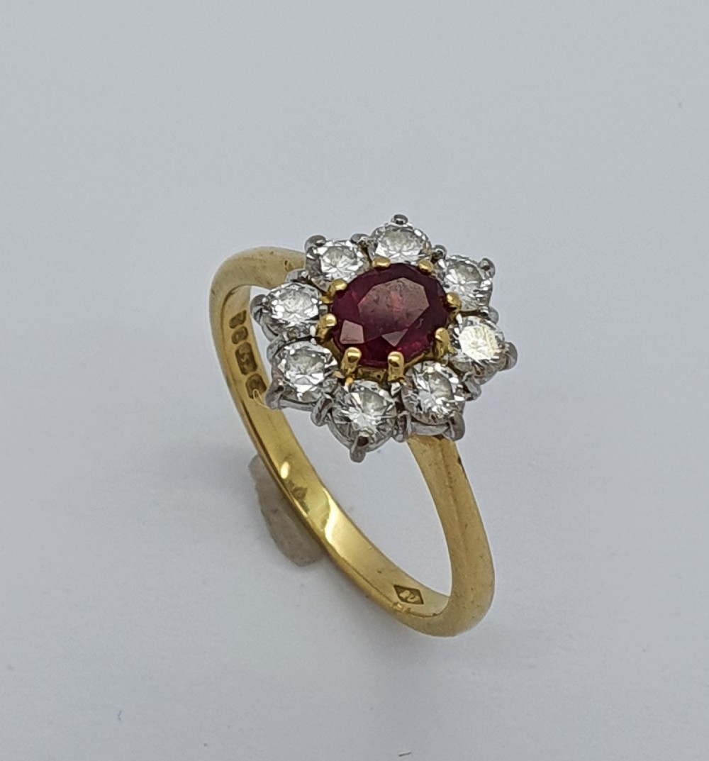 A yellow gold ring band supporting an oval red ruby framed by ten round diamonds in a claw cluster setting.