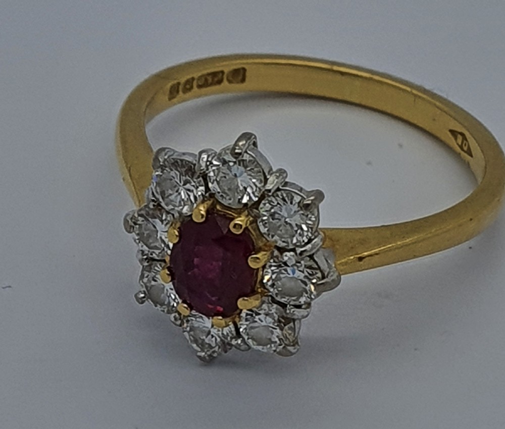 A yellow gold ring band supporting an oval red ruby framed by ten round diamonds in a claw cluster setting. View II