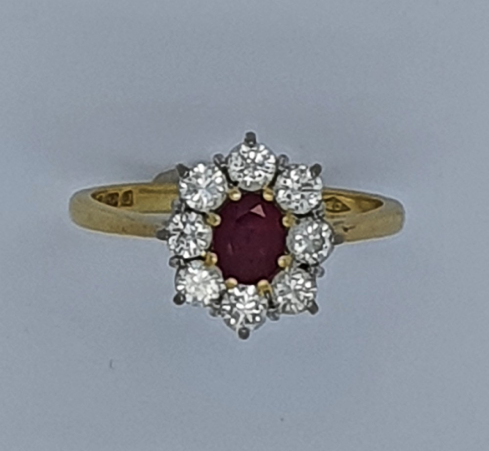 A yellow gold ring band supporting an oval red ruby framed by ten round diamonds in a claw cluster setting. View V