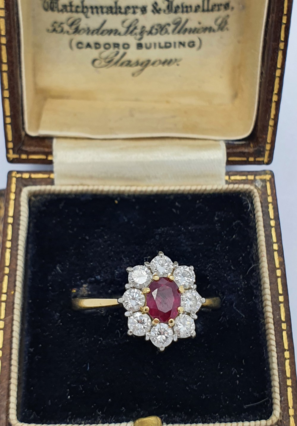 A yellow gold ring band supporting an oval red ruby framed by ten round diamonds in a claw cluster setting. View VI