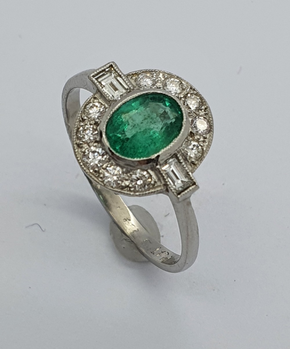 An Art Deco style white metal ring featuring a central oval green emerald within a circular diamond halo accented by baguette-cut side diamonds.