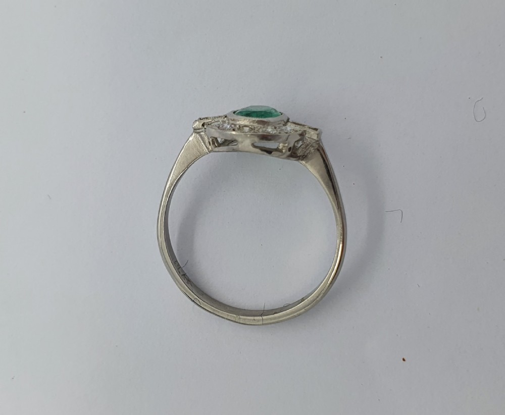 An Art Deco style white metal ring featuring a central oval green emerald within a circular diamond halo accented by baguette-cut side diamonds. View V
