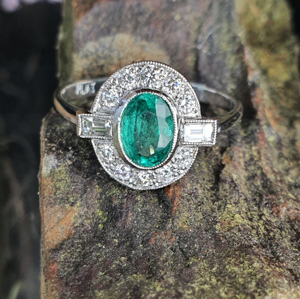 An Art Deco style white metal ring featuring a central oval green emerald within a circular diamond halo accented by baguette-cut side diamonds. View VII