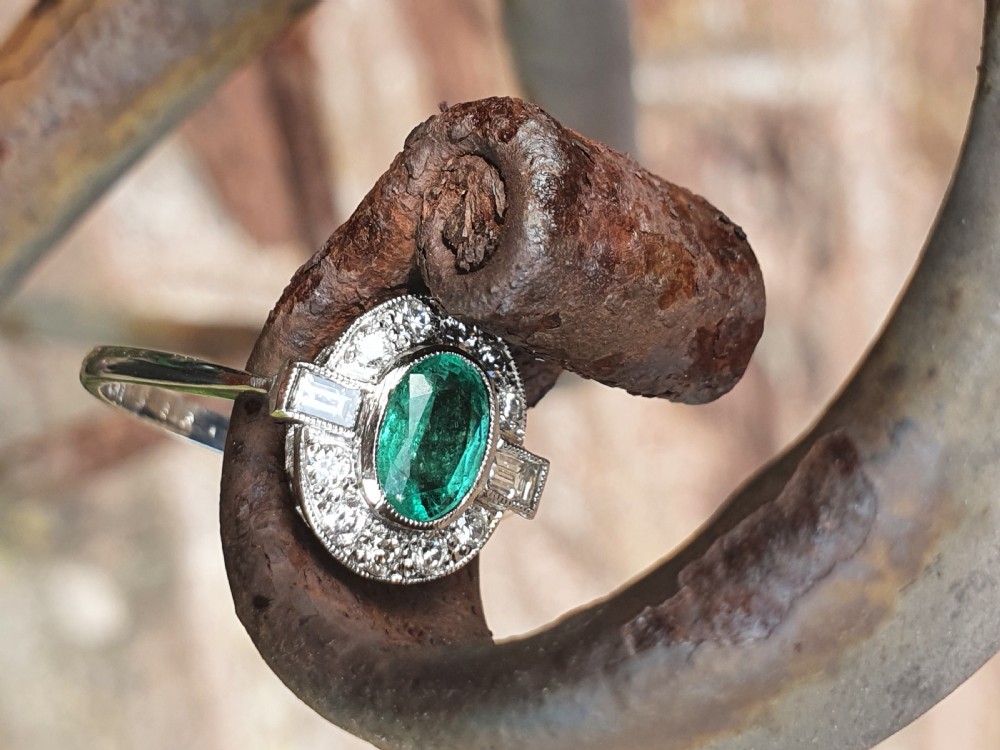 An Art Deco style white metal ring featuring a central oval green emerald within a circular diamond halo accented by baguette-cut side diamonds. View IX